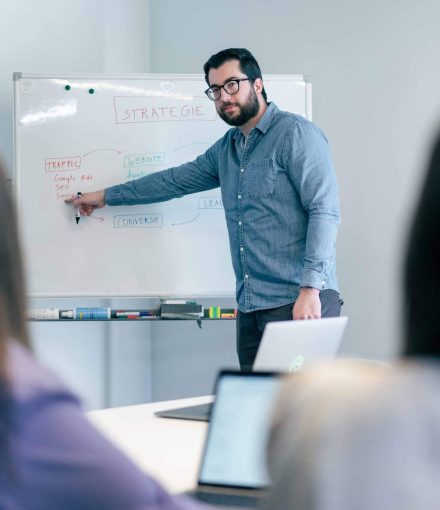 Marketingstrateeg legt online strategie uit aan team met whiteboard en laptops in vergaderruimte.